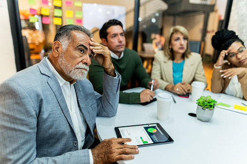 A group of professionals in a business meeting. Focus is on a man who looks like he might be struggling to hear what is being said.