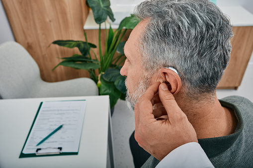 A man wearing a hearing aid has his device adjusted by an audiologist.