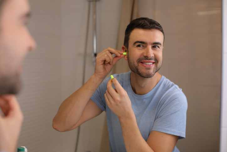 A man in his forties is using ear plugs to avoid the stress of noisy situations. He looks at his mirror reflection with positivity.