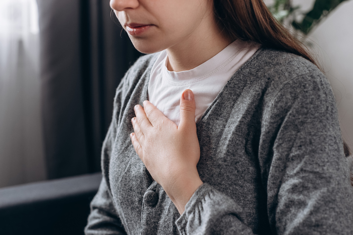 Close up of young woman feeling strong ache in chest, eyes not visible, placing hand on heart, wearing grey cardigan over white shirt.